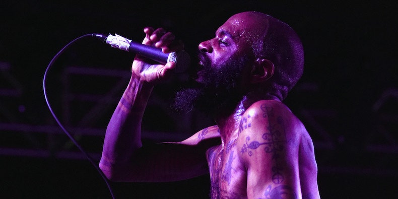 A picture of MC Ride, he is holding a microphone, and is lit by a purple light.